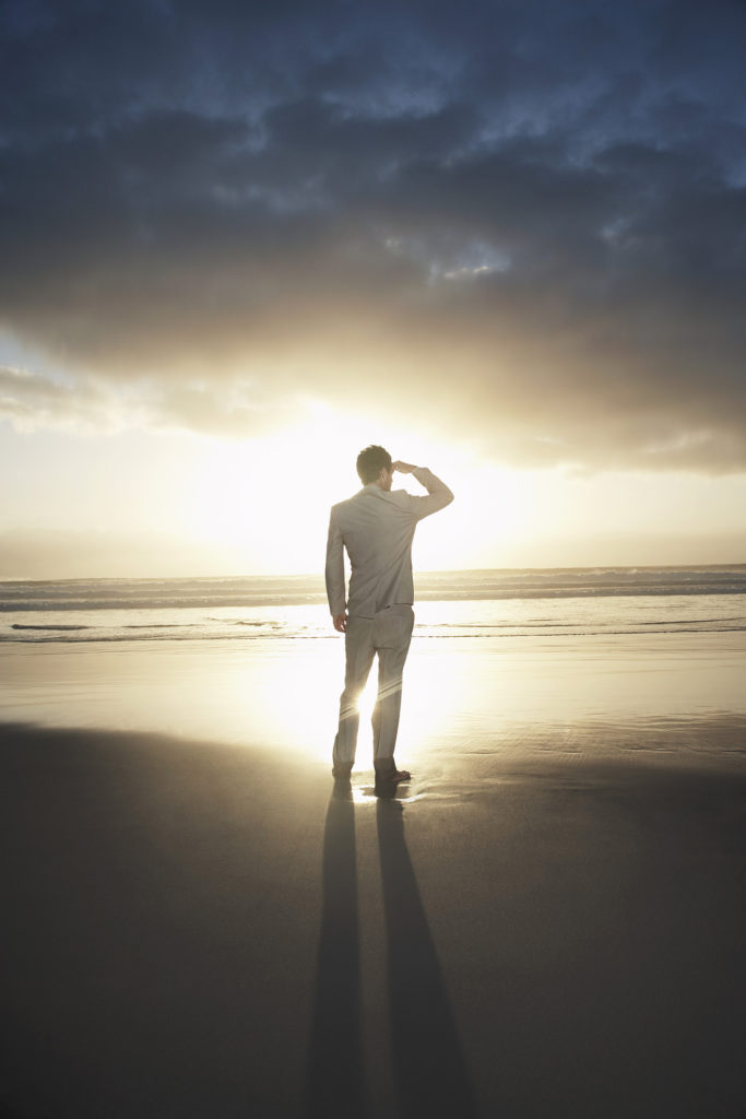 Rear view of businessman standing on beach looking to the horizon at sunset
